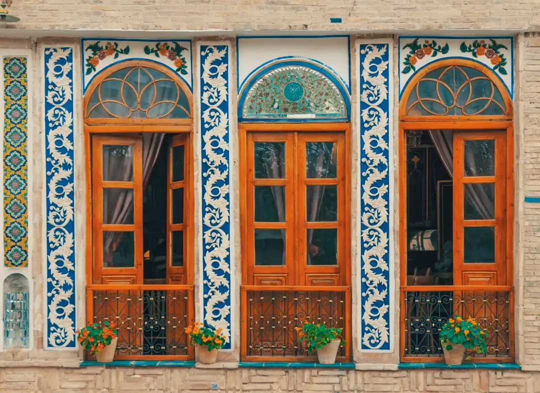 A traditional Iranian facade — arched wooden doors framed by blue and white floral stucco, with potted nasturtiums along the sill.