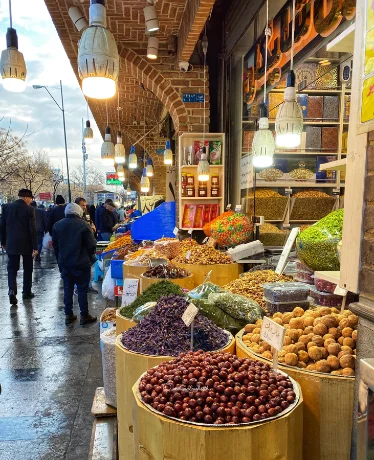 A neighbourhood bazaar storefront in winter, its open bins filled with dried fruit, nuts and pulses as shoppers pass.
